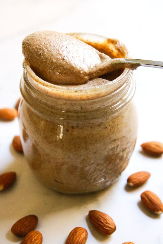 This is a vertical image looking at a glass jar filled with almond butter. A spoon is scooping almond butter and laying across the top opening of the jar. The jar sits on a white surface with almonds scattered around it.