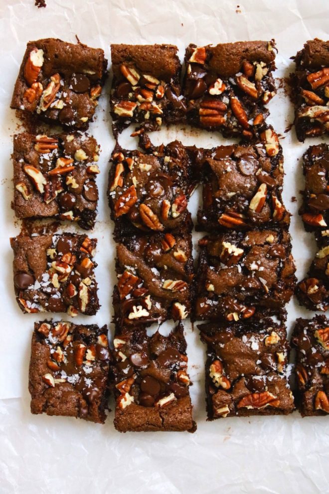 This is an overhead image looking down onto gooey chocolate almond butter brownies cut into squares and topped with pecans, chocolate and flakey salt. The brownies sit on a white piece of parchment paper.