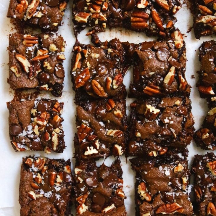 This is an overhead image looking down onto gooey chocolate brownies cut into squares and topped with pecans, chocolate and flakey salt. The brownies sit on a white piece of parchment paper.