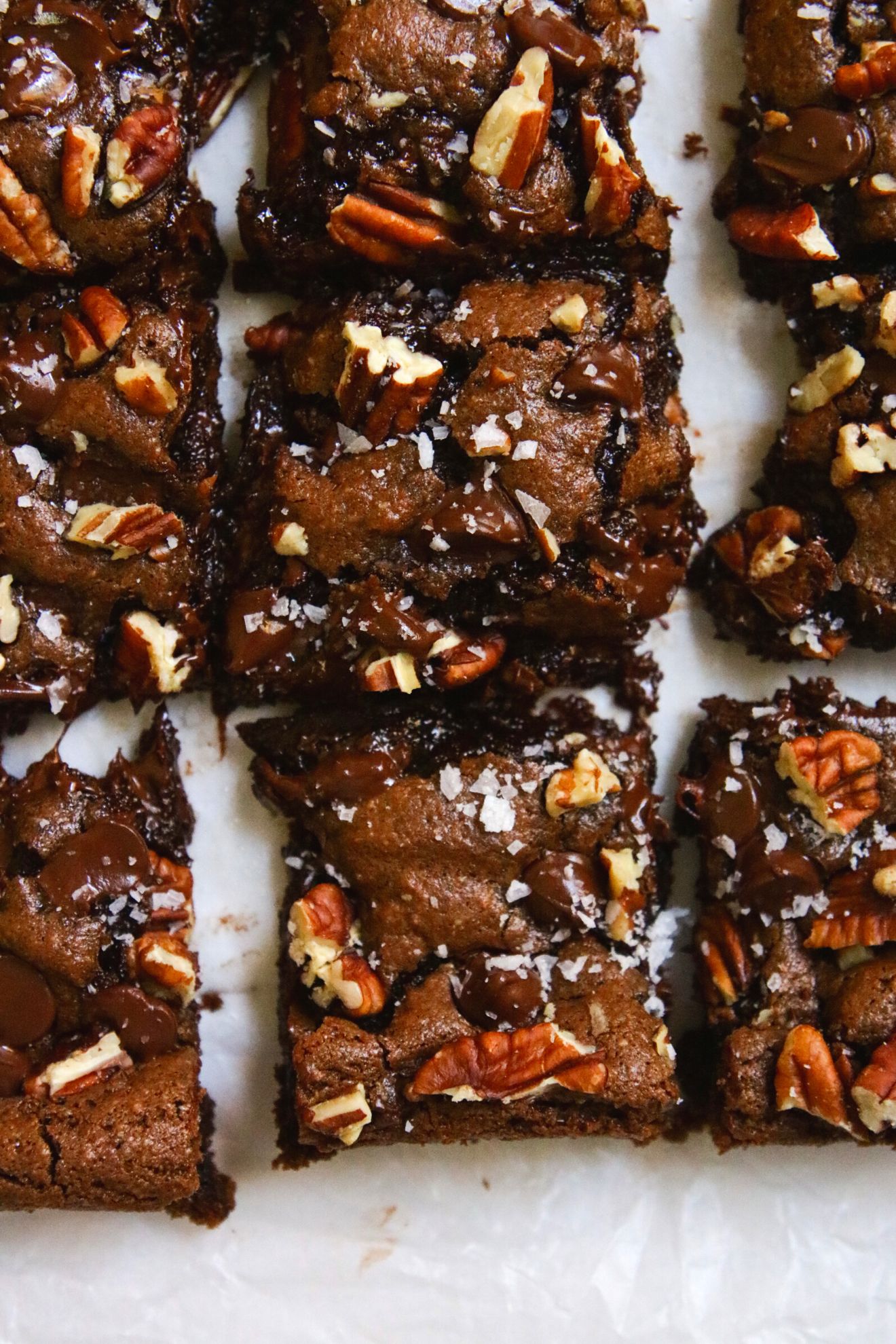 This is an overhead image of gooey brownies cut into squares. The brownies are topped with pecans and flakey salt. They sit on a white surface.