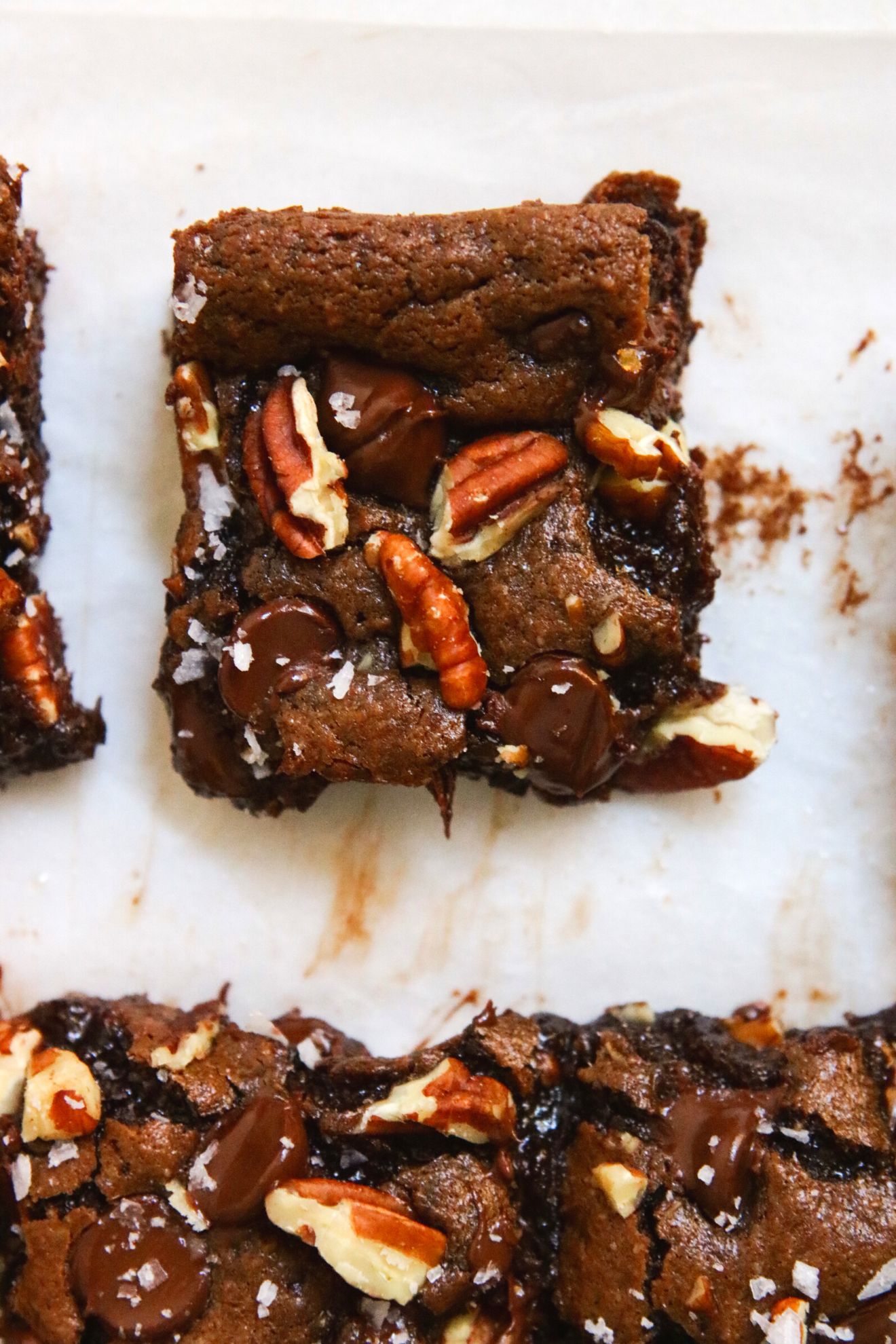 This is an overhead image of a gooey almond butter brownie topped with pecans and flakey salt. The brownie sits on a white surface with more brownies to the bottom of the image.