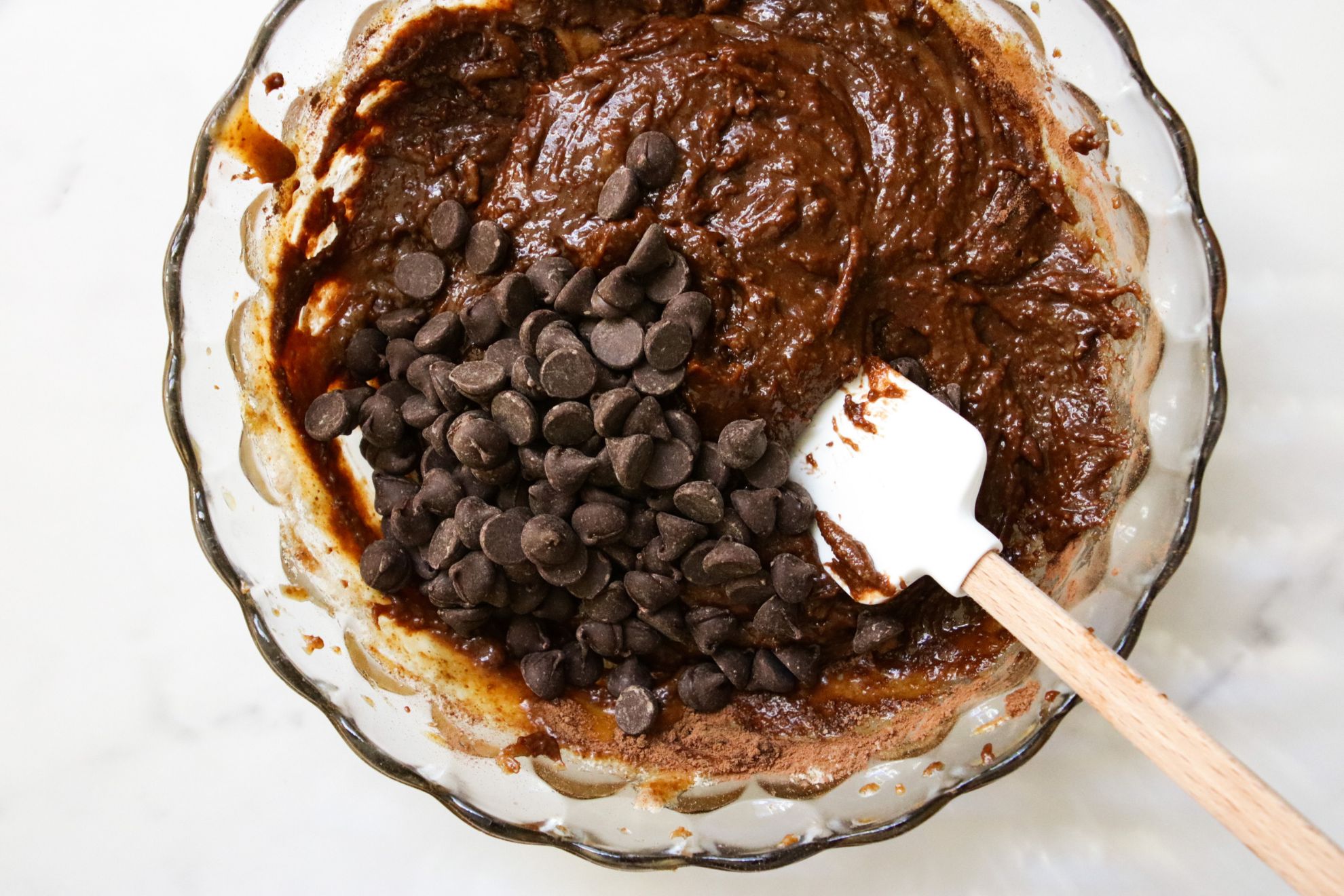 This is an overhead image looking into a glass bowl with brownie batter. There are chocolate chips on top of the brownie batter and a white rubber spatula with a wood handle in the batter and leaning on the side of the bowl. The bowl sits on a white marble counter.