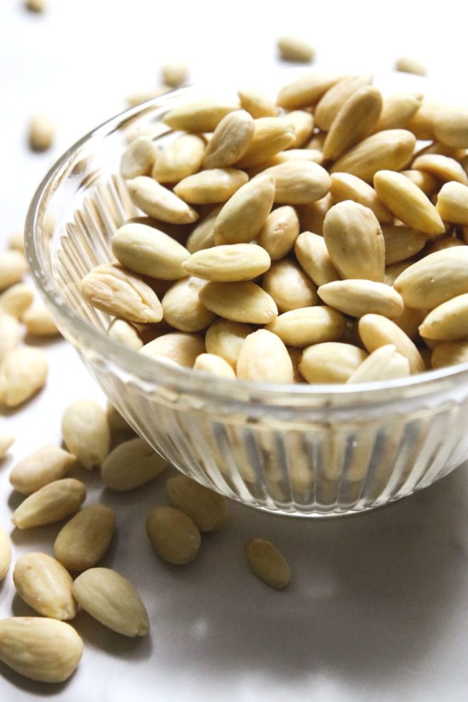 This is a side view of a glass bowl filled with blanched whole almonds. The bowl sits on a white counter and more almonds are scattered around the bowl.
