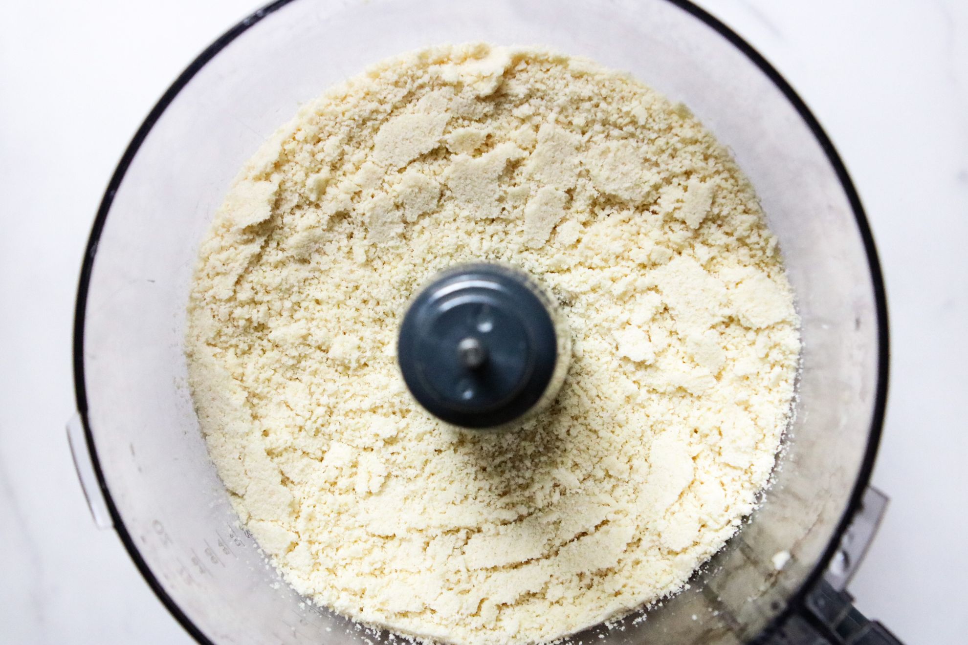 This is an overhead horizontal image of a food processor with blanched almond flour in it. The food processor sits on a white surface.