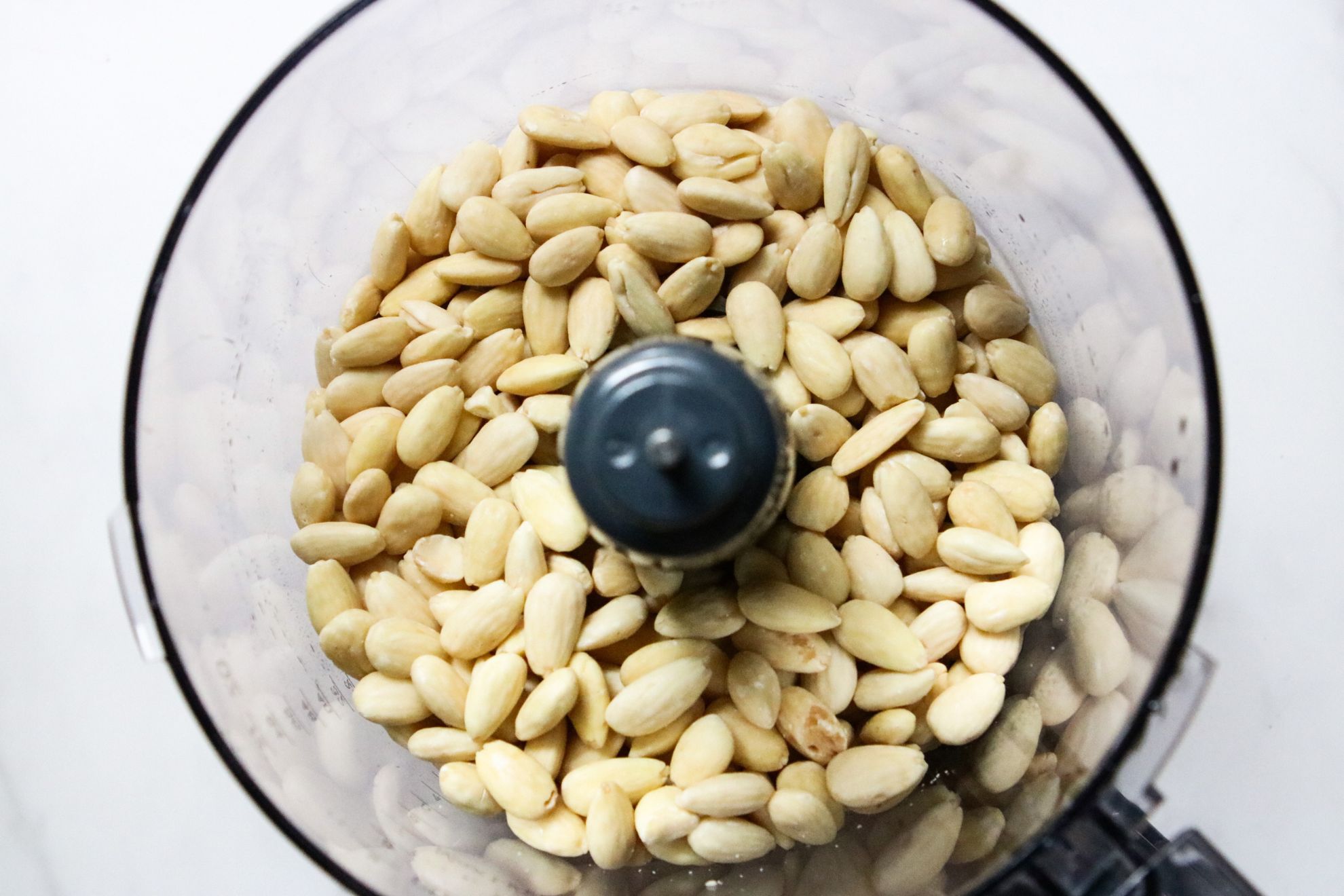 This is an overhead horizontal image of a food processor with blanched almonds in it. The food processor sits on a white counter.