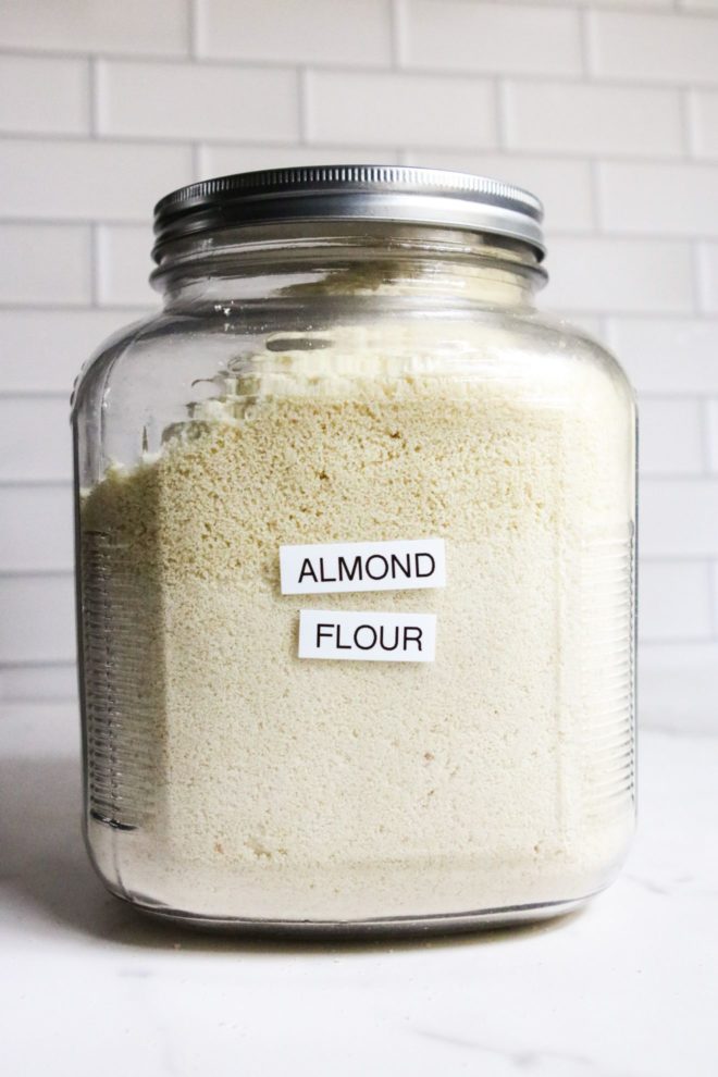 This is a side view of a large glass jar filled with blanched almond flour. The jar has a printed label that reads "almond flour" and a metal lid. The jar sits on a white marble counter with white subway tile behind it.