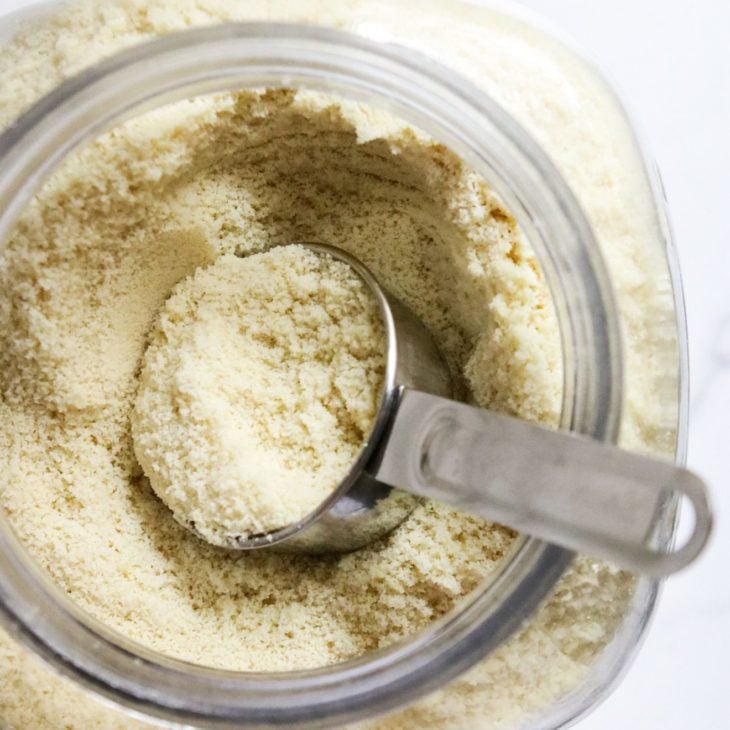This is an overhead vertical image of a large glass container with light tan flour in it. A stainless steel measuring cup is in the jar with some flour filling it. The jar sits on a white counter.