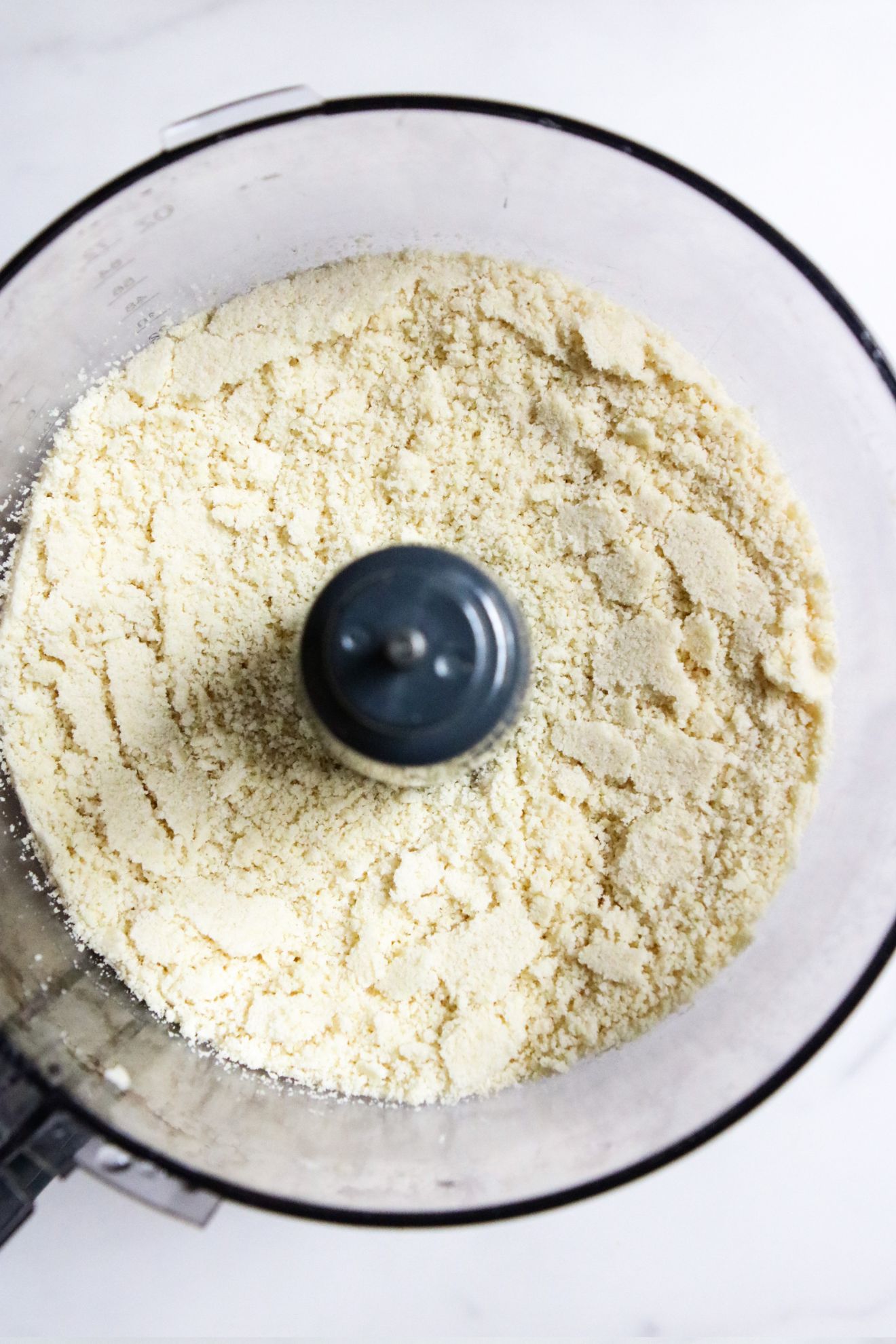 This is an overhead vertical image of a food processor with blanched almond flour in it. The food processor sits on a white counter.