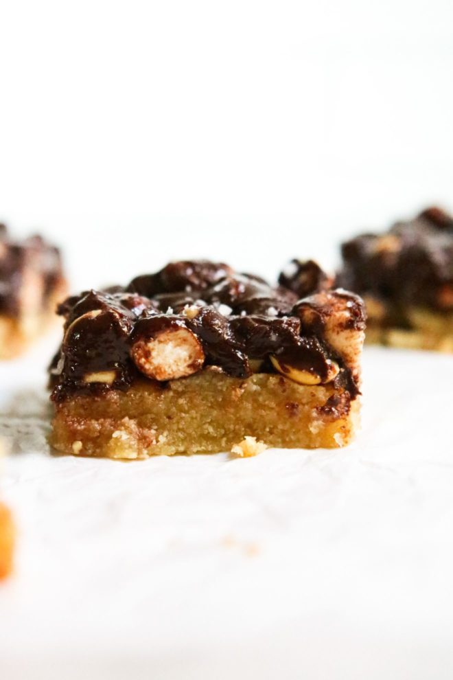 This is a vertical image of a side view of an almond flour shortbread rocky road bar. The square has a shortbread crust and is topped with chocolate, mini marshmallows and peanuts. The square sits on a white surface with a white background and more bars blurred in the background.