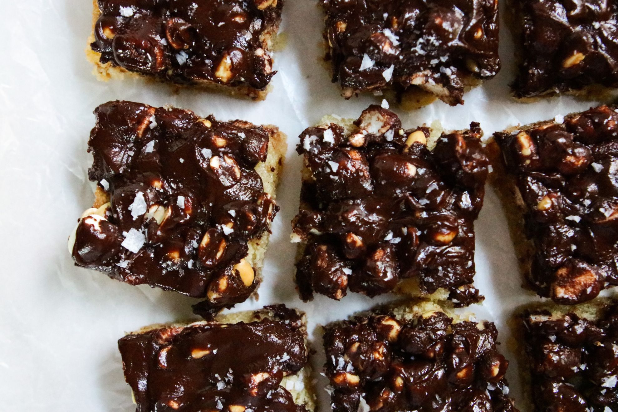 This is a horizontal overhead image of shortbread and chunky chocolate bars cut into squares. The bars sit on a white piece of parchment paper and sprinkled with flakey salt.
