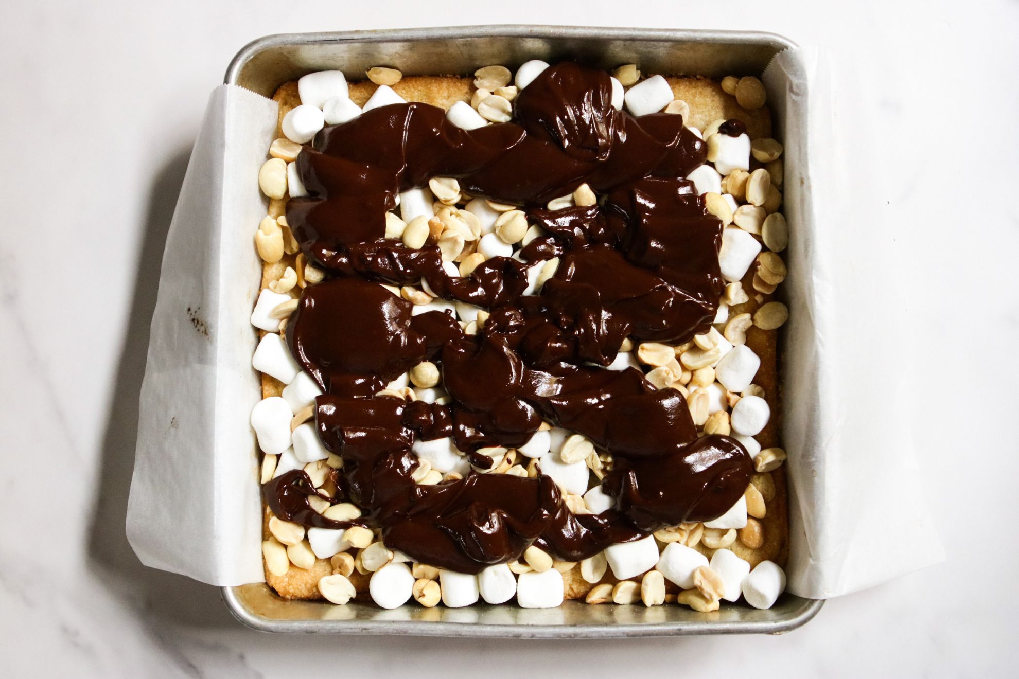 This is an overhead horizontal image of a square pan lined with parchment paprt. It has a shortbread crust with mini marshmallows, peanuts, and chocolate drizzled on top. The square sits on a white marble counter.