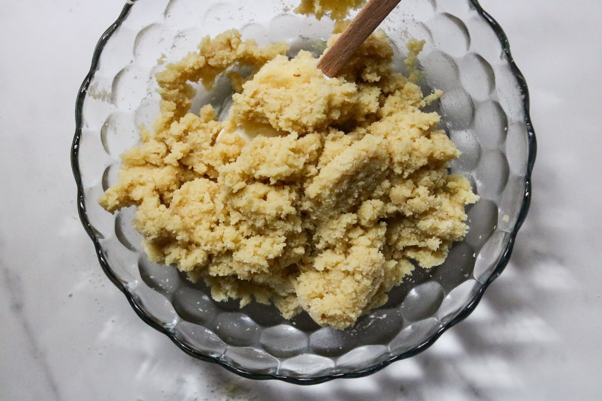 This is a horizontal overhead image of a glass bowl with a raw shortbread mixture. A spatula is in the bowl with the mixture. The bowl sits on a white marble counter.