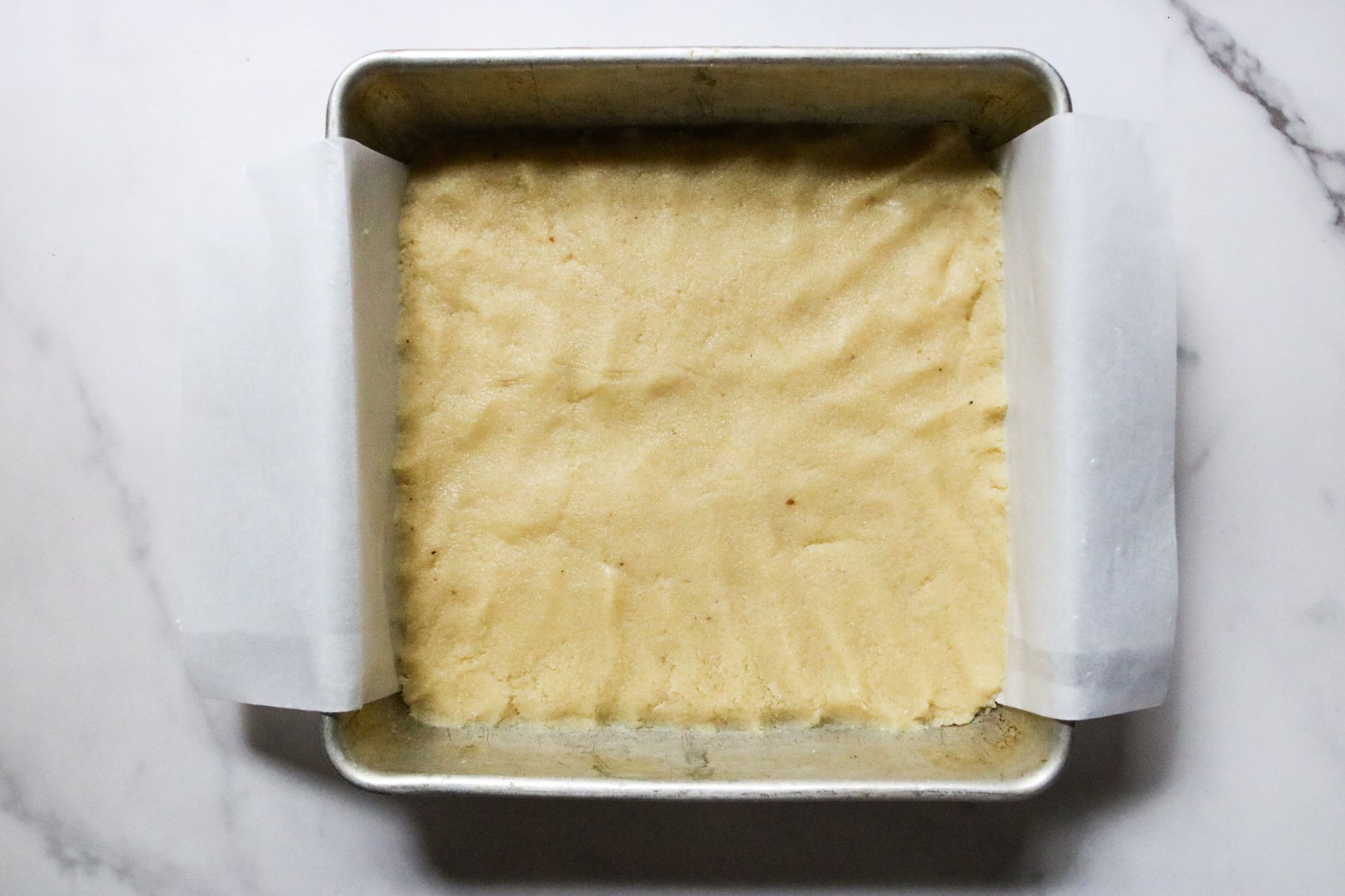 This is an overhead horizontal image of a square pan with parchment paper and a raw shortbread crust. The pan sits on a white marble counter.