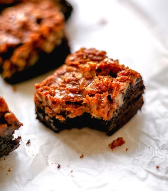 This is a side view of a brownie with a pecan pie topping and a bite taken out of it. The brownie sits on a white piece of parchment paper with more brownies blurred in the background.