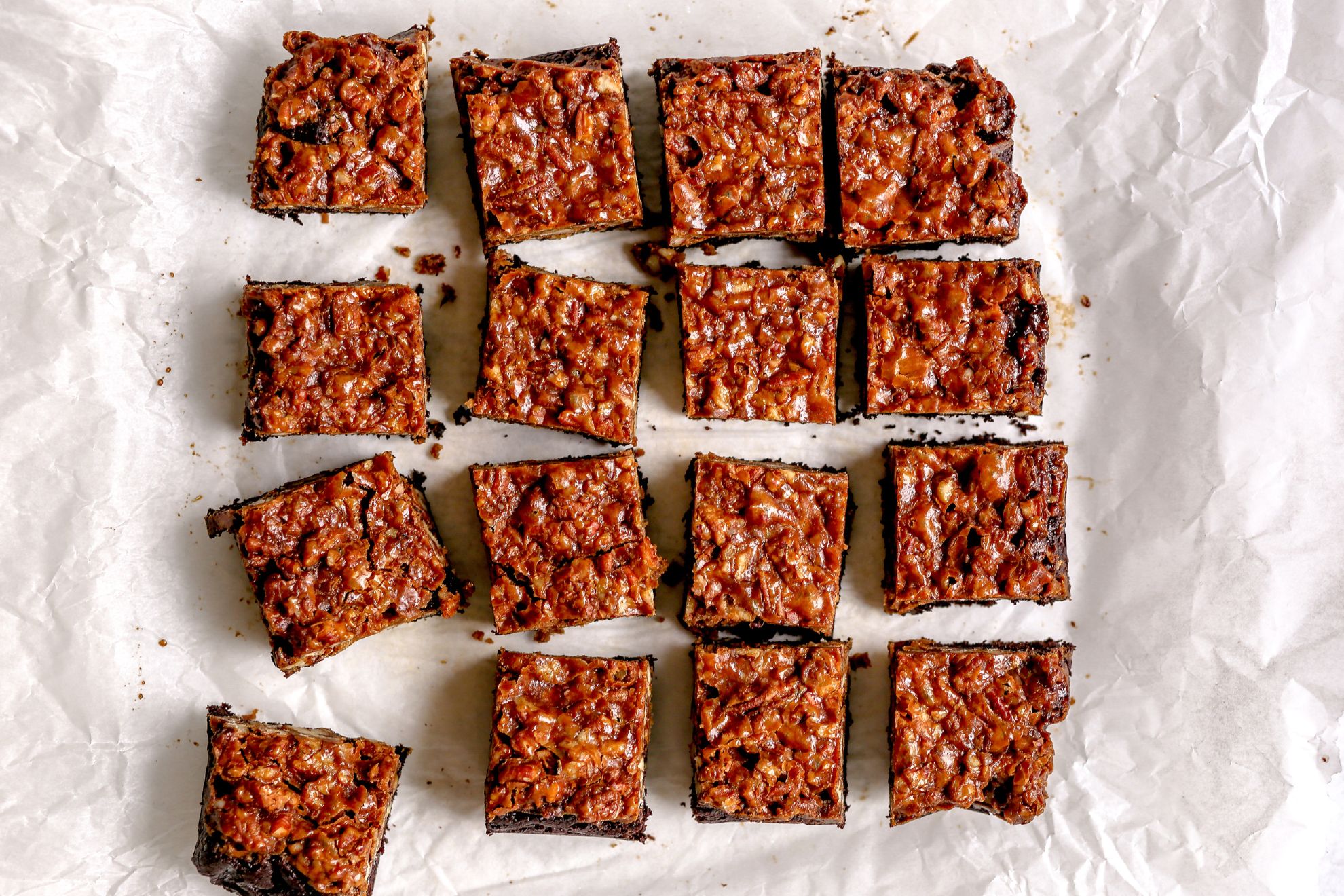 This is an overhead image of pecan pie brownies cut into 16 squares. The brownies are on a white piece of parchment paper.