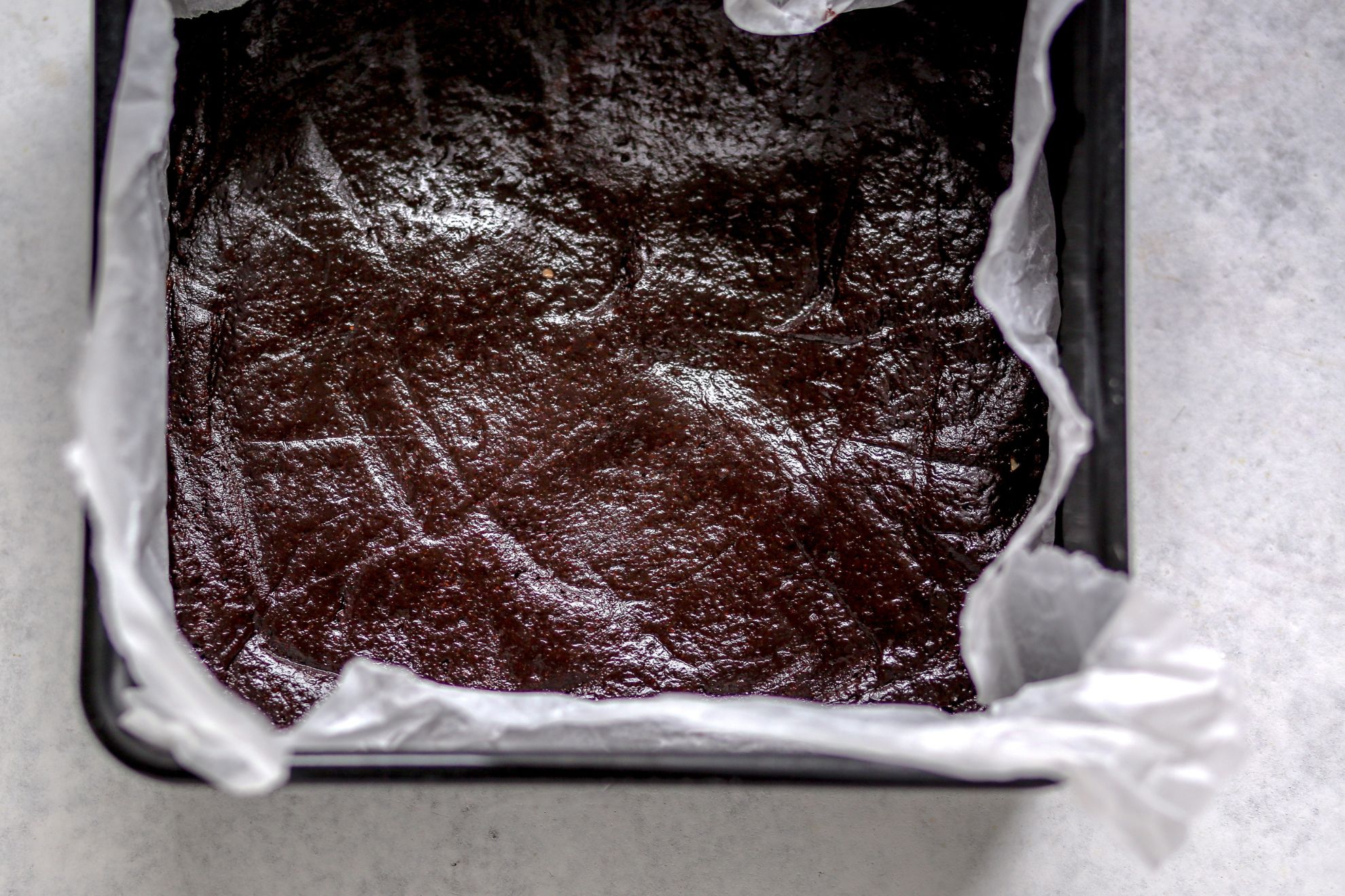 This is an overhead image of a square pan lined with parchment paper and filled with a fudgey raw brownie batter. The pan sits on a light grey surface.