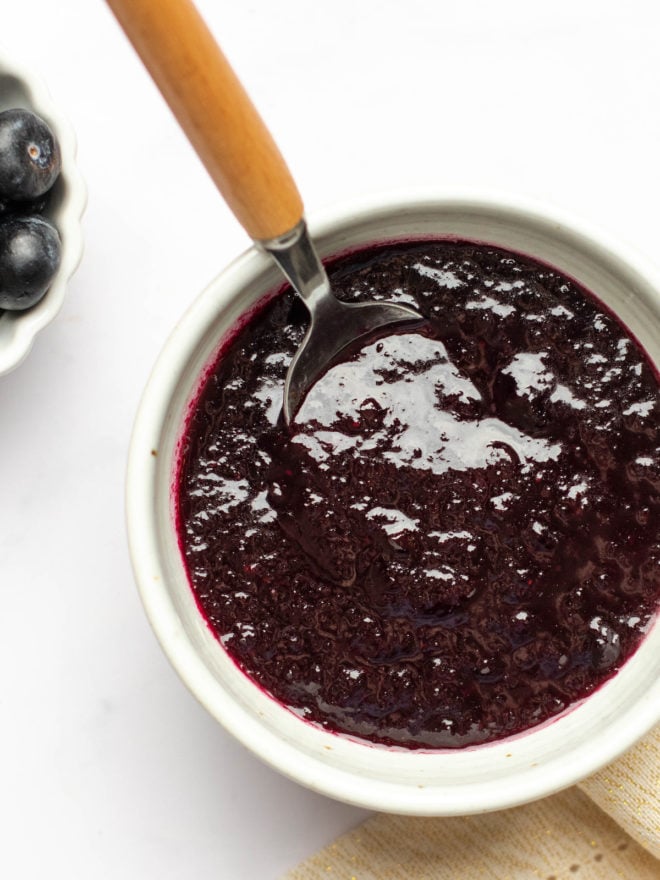 This is an overhead image of a white bowl filled with blueberry glaze. The bowl sits on a white surface and a spoon with a wooden handle is dipping into the bowl and leaning against the side. Part of a bowl of blueberries is to the top left of the image.