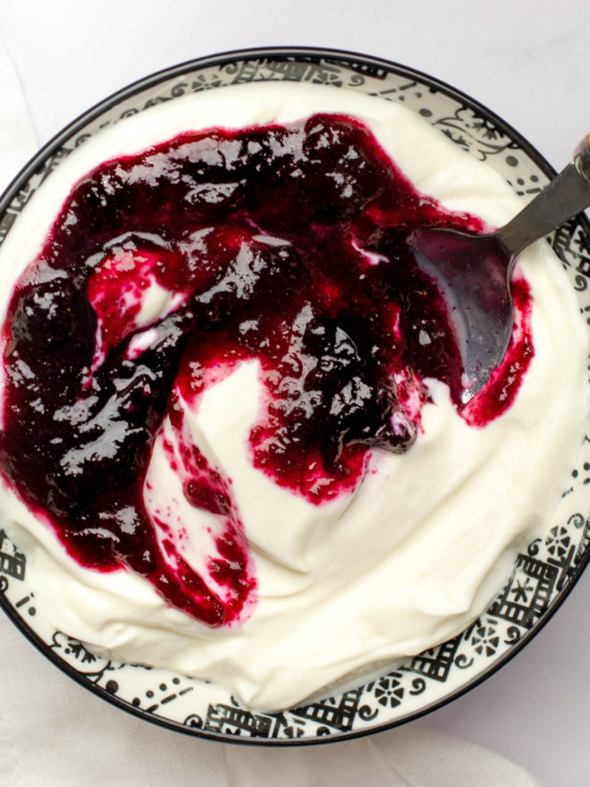 This is an overhead image of a bowl filled with yogurt and topped with blueberry glaze. A spoon is dipping into the yogurt and leaning against the side of the bowl. The bowl sits on a white surface.