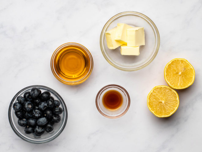 This is an overhead image of small glass bowls filled with ingredients like butter and blueberries. The bowls sit on a white marble surface.