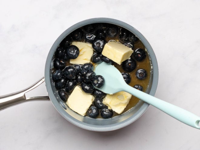 This is an overhead image of a pot with blueberries and butter. The pot sits on a white surface and a light blue spatula is leaning against the side.