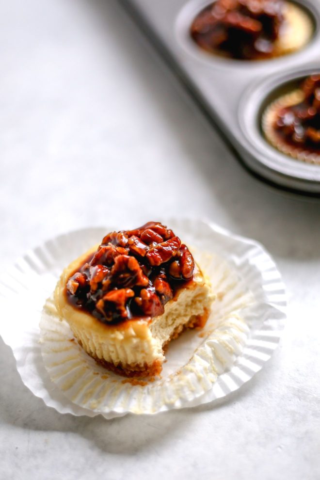 This is a side view of an individual cheesecake with a white cupcake liner pulled away and a bite taken out of it. The individual cheesecake has a caramel pecan topping and is sitting on a white surface. A muffin tin filled with more individual cheesecakes is blurred in the background.