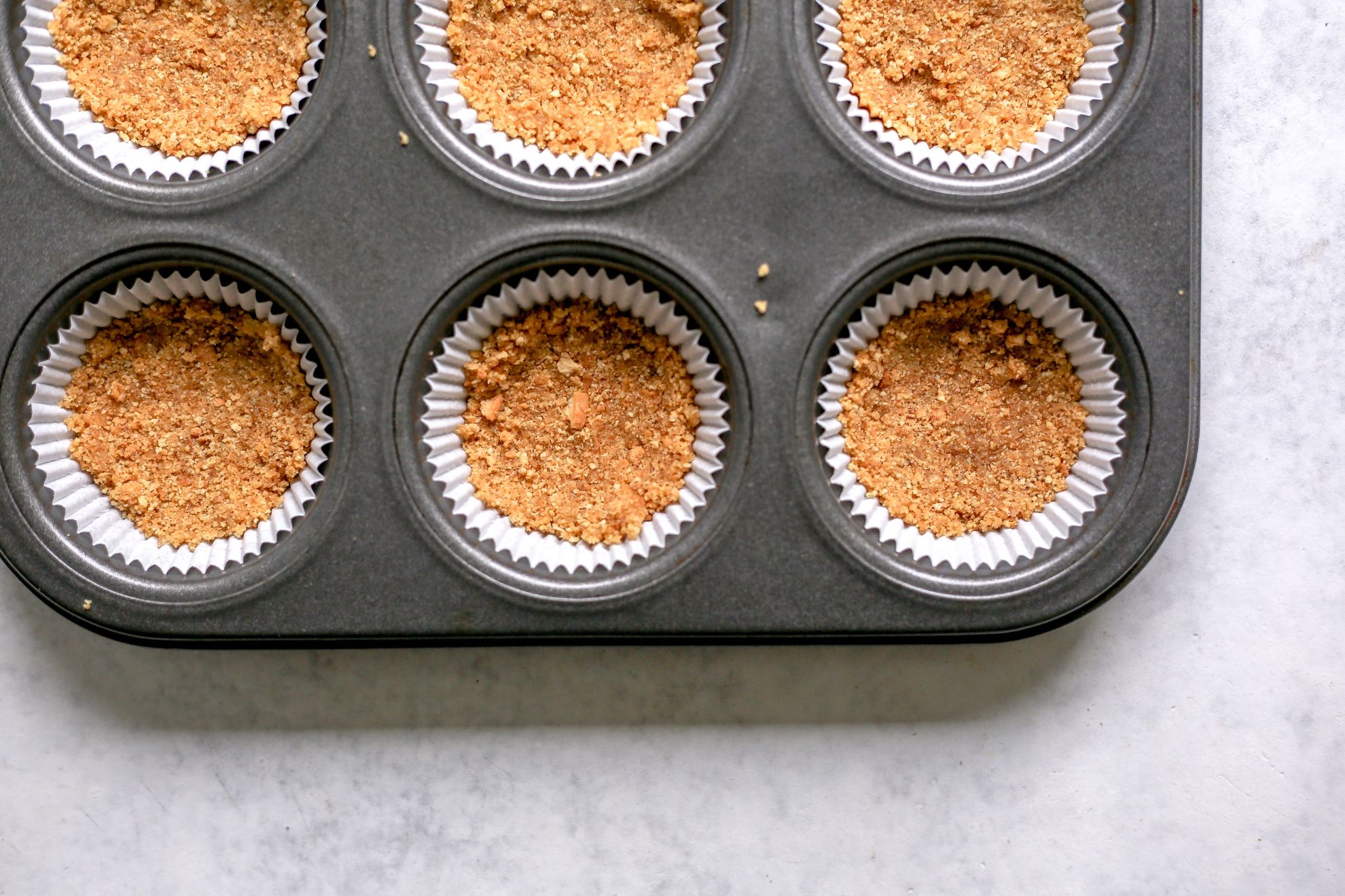 This is an overhead image of a muffin tin lined with white muffin liners and a graham cracker crust pressed into the bottom of each liner.
