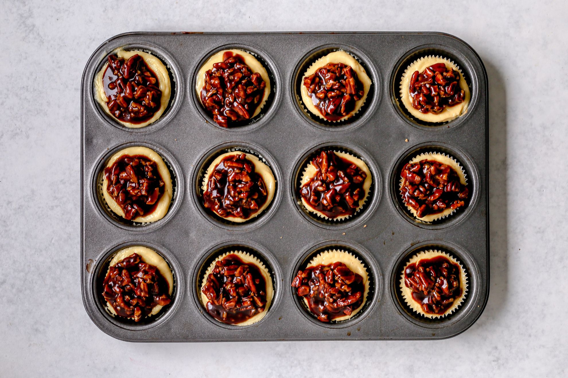 This is an overhead image of a muffin tin filled with individual cheesecakes topped with pecans. The muffin tin sits on a light surface.