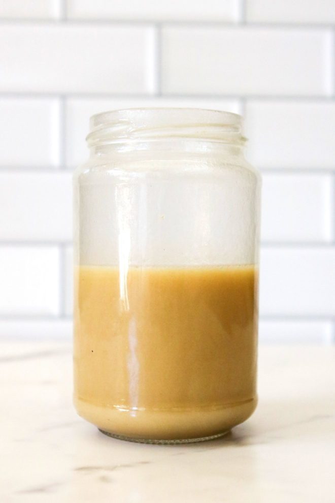 This is a side view of a jar with a lemon vinaigrette filling it halfway up. The jar sits on a marble counter with white subway tile in the background.