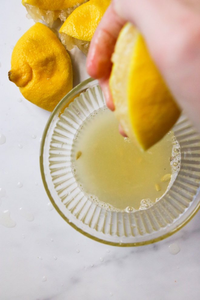 This is an image of a hand squeezing juice out of half of a lemon into a glass bowl. Squeezed lemon halves are to the top left of the image. Juice is in the glass bowl with some seeds floating in it. The bowl sits on a white marble counter.