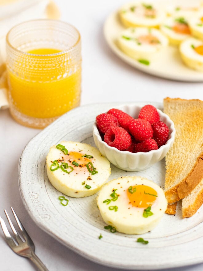 This is a vertical image of a white plate with two baked eggs, a small bowl of raspberries, and two slices of toast on it. The eggs are topped with ground pepper and scallions. The plate sits on a white surface with a fork to the bottom left of the image. Behind the plate is a glass of orange juice and to the top right of the image is a tan plate with more baked eggs.