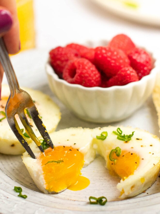 This is a vertical close up image of a baked egg cut in half The egg sits on a white plate and is topped with ground pepper and green scallion. The egg yolk is dripping out of the left half of the egg as a fork pierces it. More eggs are on the plate along with a small bowl of raspberries. Blurred in the background you can see a small glass of orange juice.