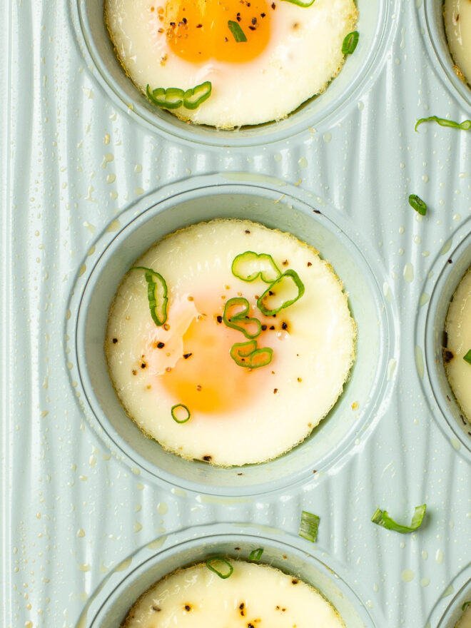 This is an overhead vertical image of a light blue muffin tin filled with baked eggs. The image is a close up of one muffin tin cavity. The eggs are topped with ground pepper and chopped green scallion. The muffin tin sits on top a white and yellow dish towel.