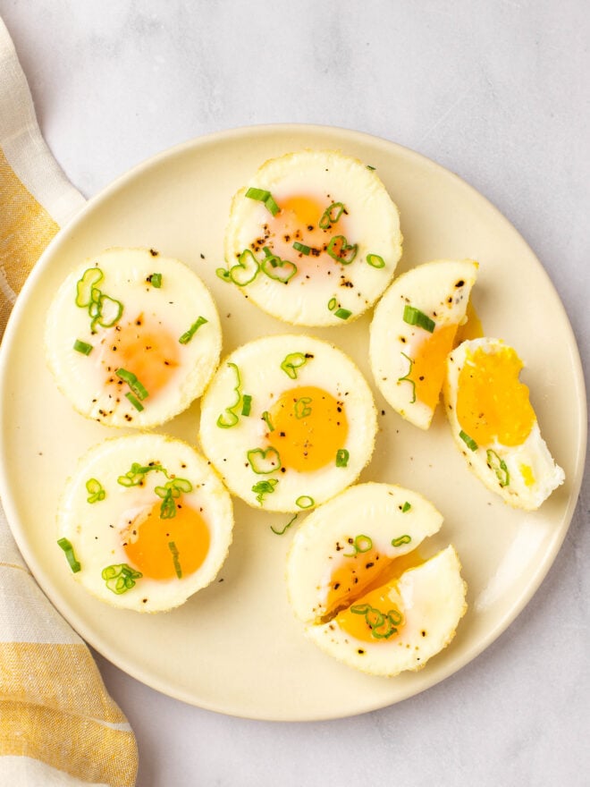 This is an overhead vertical image of a tan plate with baked eggs on top it. The eggs are in a circle and two of them are cut in half. The eggs are topped with ground pepper and chopped green scallions. The plate sits on a white marble counter with a white and yellow dishtowel to the left of the image.