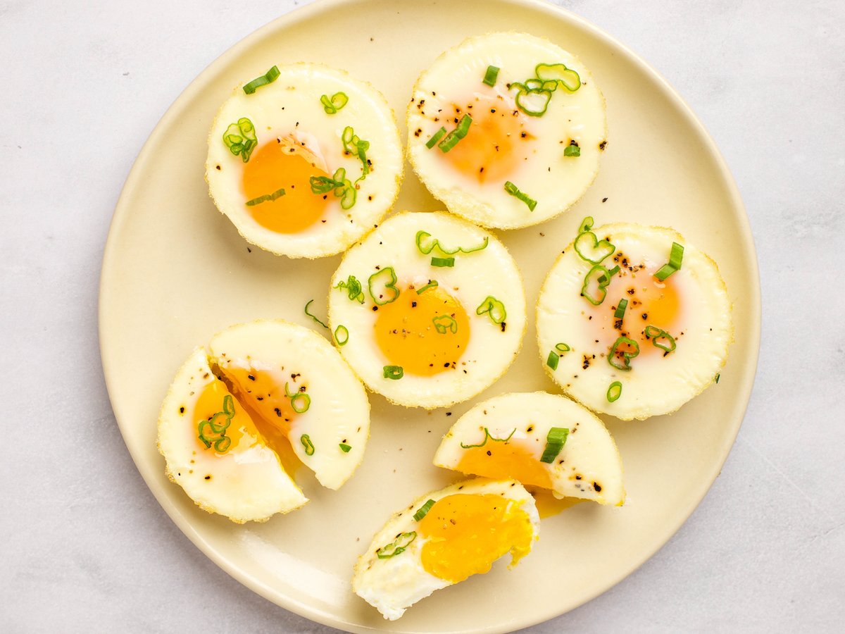 This is an overhead horizontal image of a tan plate with baked eggs on top it. The eggs are in a circle and two of them are cut in half. The eggs are topped with ground pepper and chopped green scallions. The plate sits on a white marble counter.