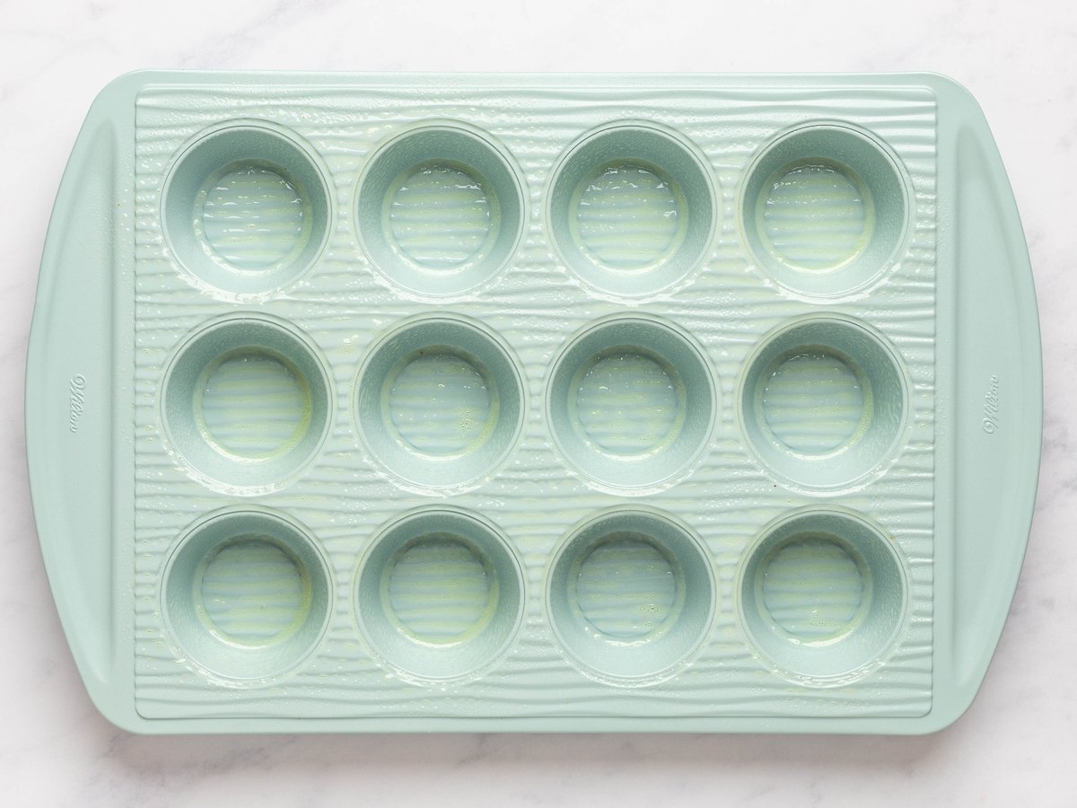 This is an overhead horizontal image of a light blue muffin tin sprayed with cooking oil. The blue muffin tin sits on a white marble counter.
