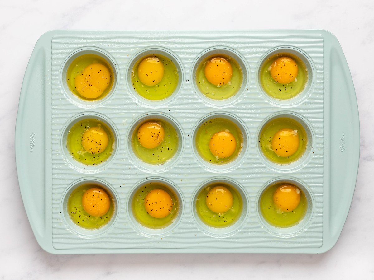 This is an overhead horizontal image of a light blue muffin tin sprayed with cooking oil. The blue muffin tin sits on a white marble counter. Each muffin cavity has a raw egg cracked in it and sprinkled with visible specs of pepper.