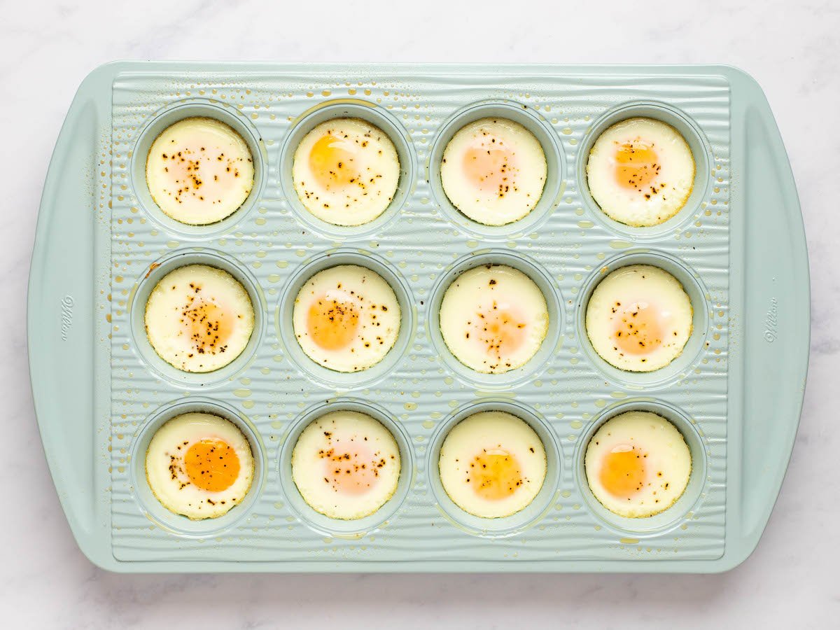 This is an overhead horizontal image of a light blue muffin tin sprayed with cooking oil. The blue muffin tin sits on a white marble counter. Each muffin cavity has a baked egg in it and sprinkled with visible specs of pepper.