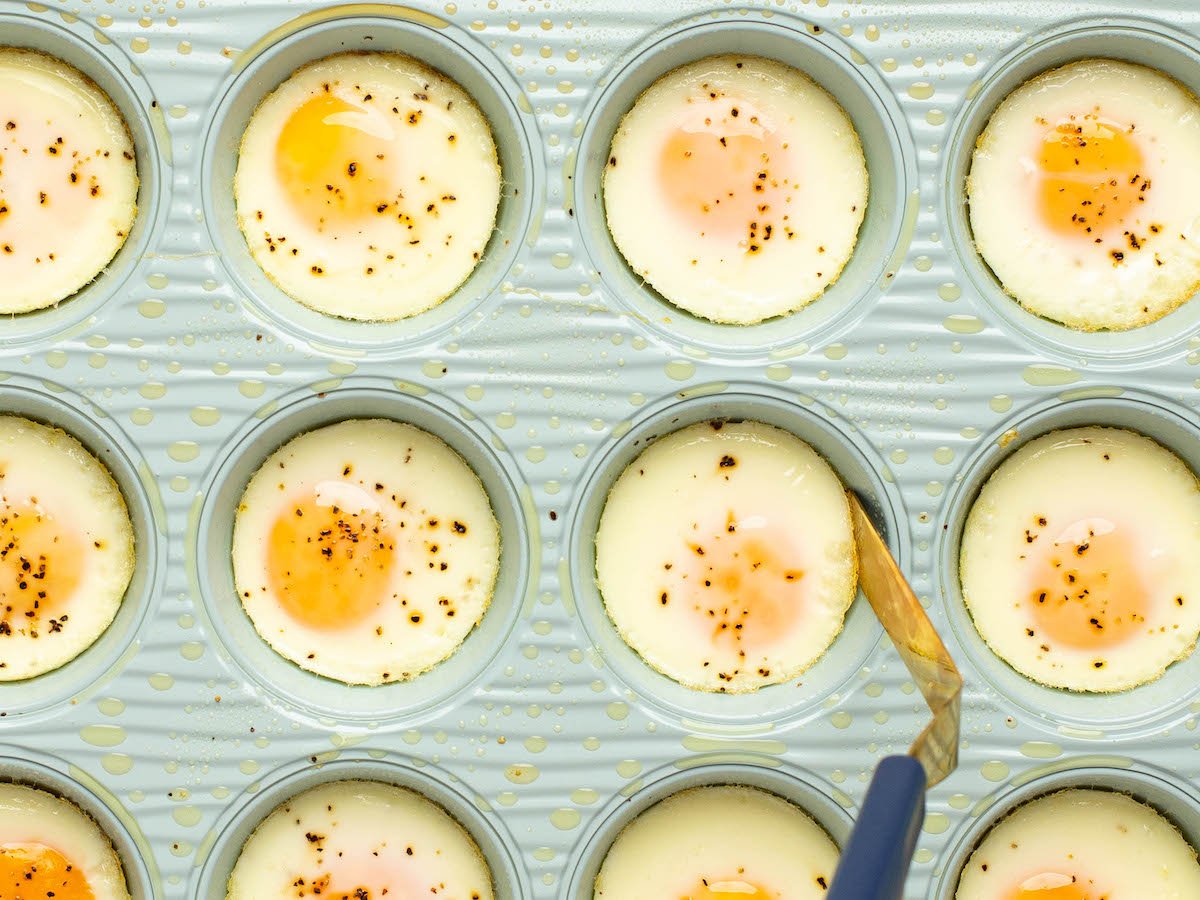 This is an overhead horizontal image of a light blue muffin tin sprayed with cooking oil. Each muffin cavity has a baked egg in it and sprinkled with visible specs of pepper. A small spatula is sliding under one of the middle eggs to remove it from the tin.