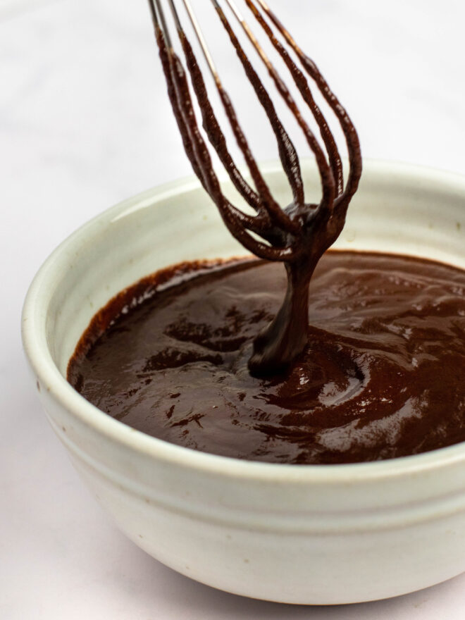 This is a vertical image of an angled view of a white bowl filled 3/4 the way with chocolate glaze. A whisk is coming from the top of the image, coated with chocolate sauce and streaming some down into the bowl. The bowl sits on a white surface.