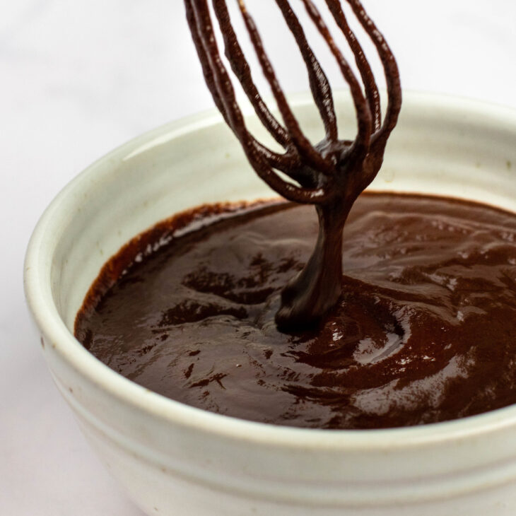 This is a vertical image of an angled view of a white bowl filled 3/4 the way with chocolate glaze. A whisk is coming from the top of the image, coated with chocolate sauce and streaming some down into the bowl. The bowl sits on a white surface.