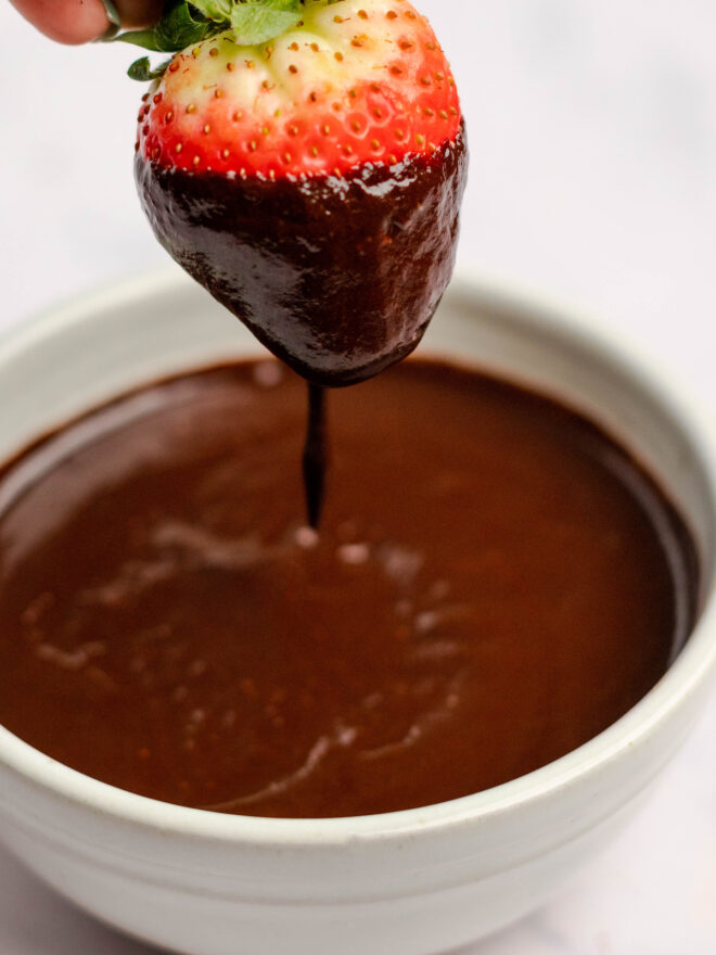 This is a vertical image of a side view of a white bowl filled with chocolate glaze. A strawberry is being held at the top of the image dripping in chocolate glaze. The white bowl sits on a light surface.