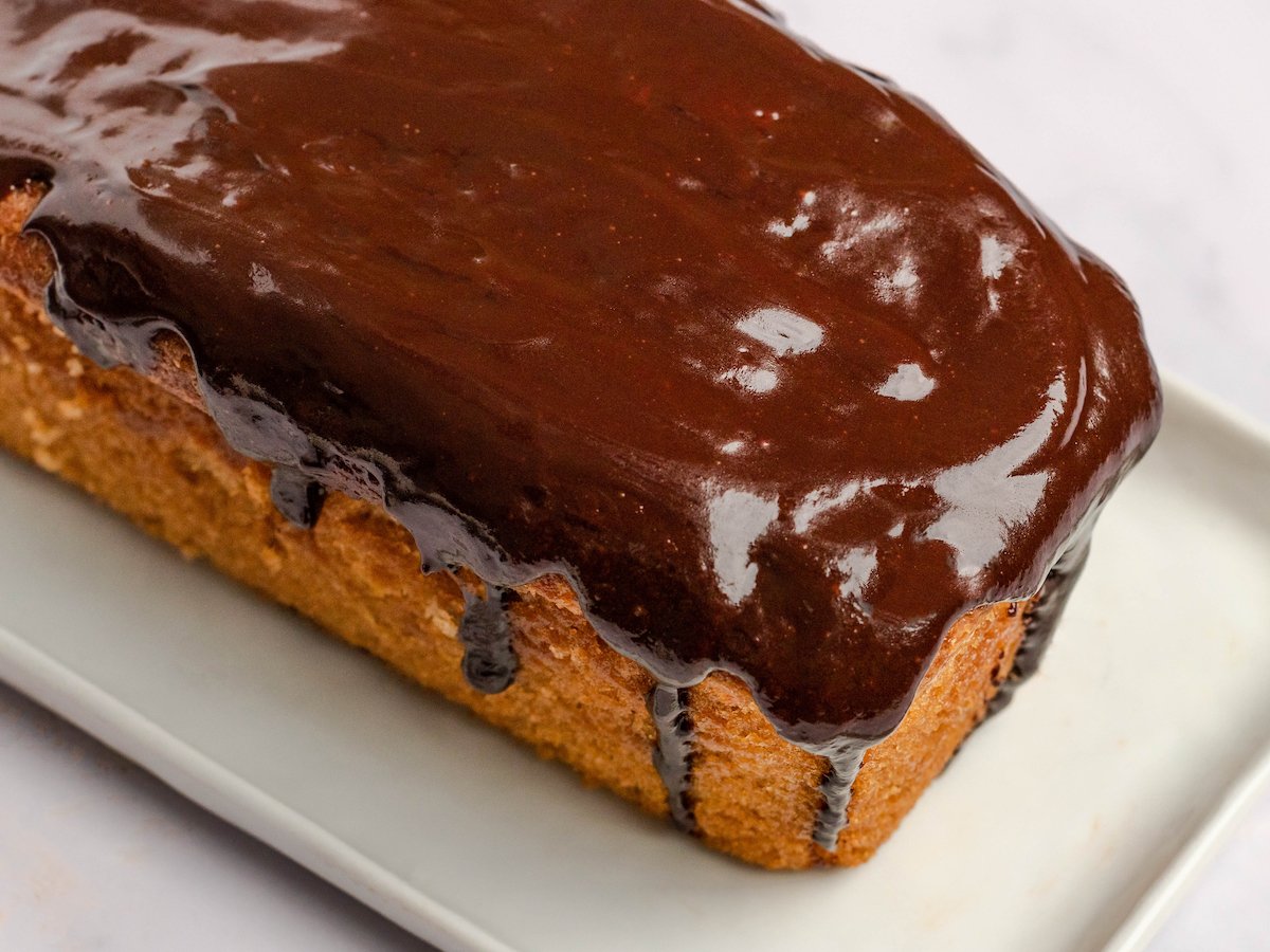 This is a horizontal image of a loaf cake on a white rectangle plate. The cake is covered in a chocolate glaze with some dripping down the sides. The rectangle plate sits on a white surface. 