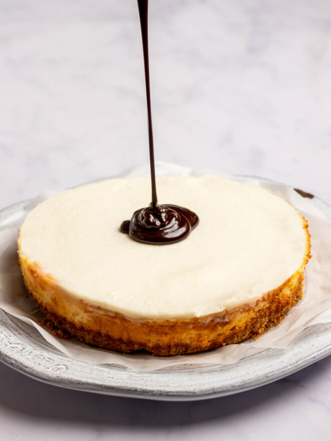 This is a vertical image of a side view of a cheesecake with a chocolate glaze being drizzle into the center of the cake and a stream of chocolate coming from the top of the image. The cake sits on a piece of white parchment paper on a white plate on a light marble surface.