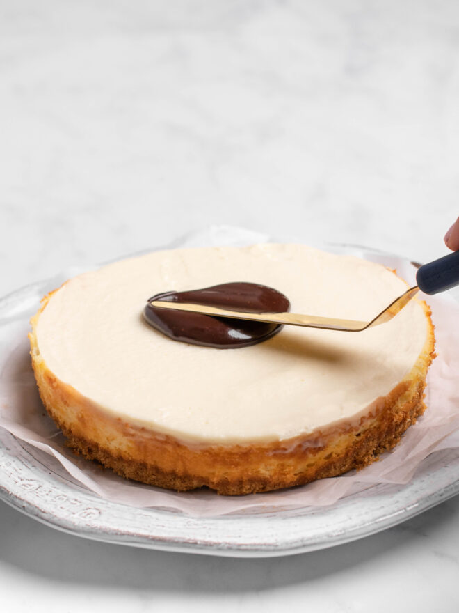 This is a vertical image of a side view of a cheesecake with a chocolate glaze being spread across the center of the cake. The cake sits on a piece of white parchment paper on a white plate on a light marble surface.