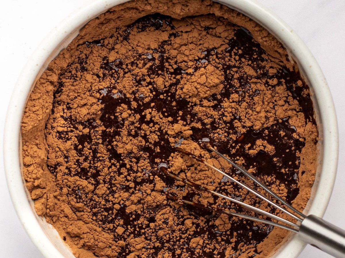 This is an overhead image of a white bowl filled with chocolate liquid and cocoa powder. A whisk is in the chocolate, leaning against the side of the bowl with the handle of the whisk to the bottom right corner of the image. The bowl sits on a light marble surface.