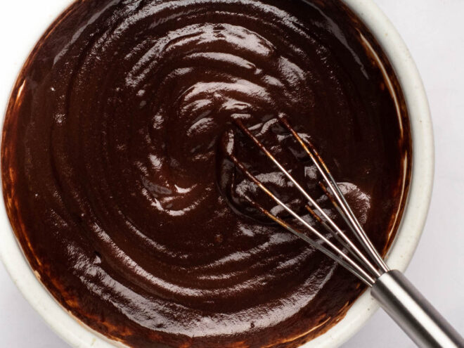 This is an overhead image of a white bowl filled with chocolate glaze. A whisk is in the chocolate, leaning against the side of the bowl with the handle of the whisk to the bottom right corner of the image. The bowl sits on a light marble surface.