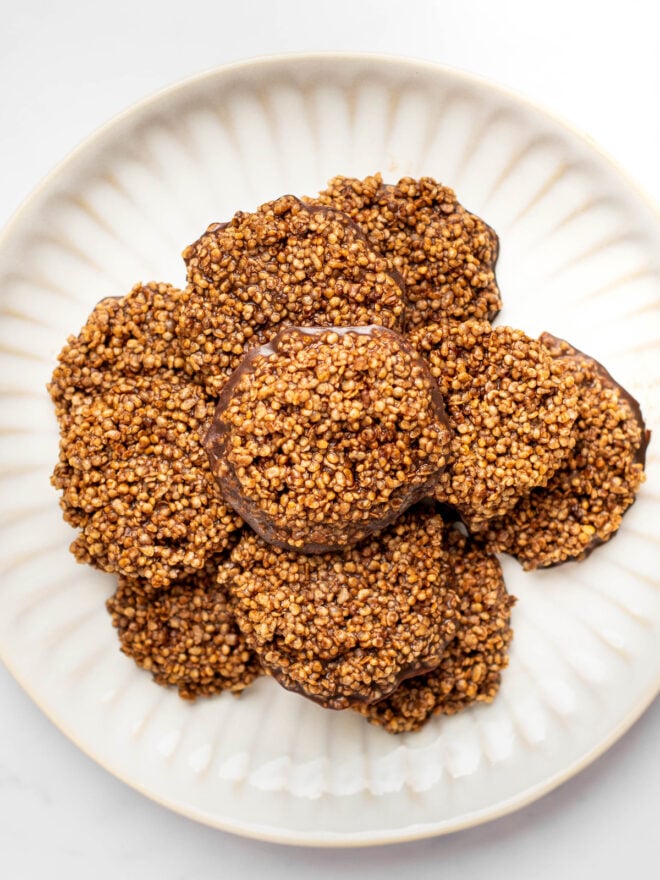 This is an overhead vertical image of chocolate quinoa crisps in a circular cookie shape. The stack of chocolate crisps is stacked on a white plate on a white marble surface.