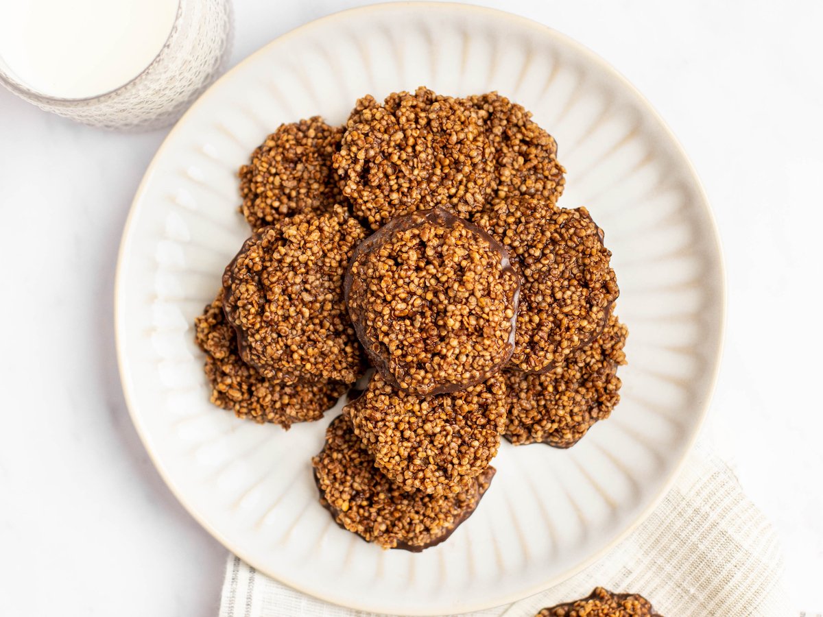 This is an overhead image of chocolate quinoa crisps in a circular cookie shape. The stack of chocolate crisps is stacked on a white plate on a white marble surface. A small glass of milk is to the top left corner of the image. To the bottom right of the image is a tan textured dish towel with a part of a quinoa crisp peeking into the image from the bottom.