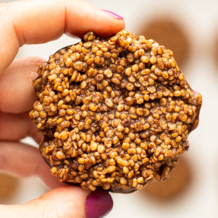 This is a horizontal image of a hand with deep pink nail polish holding a chocolate quinoa crisp in between their thumb and pointer finger. More chocolate quinoa crisps are blurred in the background.