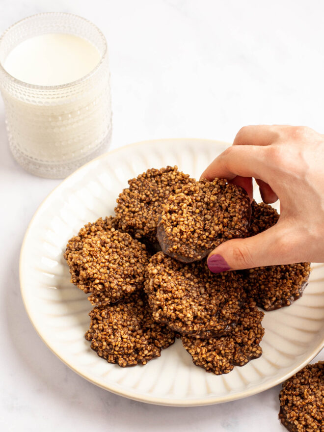 This is a vertical image viewed from an angle looking onto a white plate stacked with circle chocolate quinoa crisps. The plate sits on a white marble surface. A chocolate quinoa crisp is to the bottom right corner and a glass of milk is to the top left.